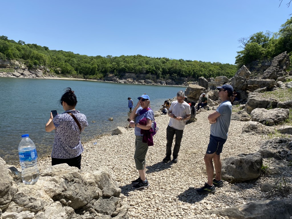 People stand along a rocky beach. Trees and small cliffs are visible in the background